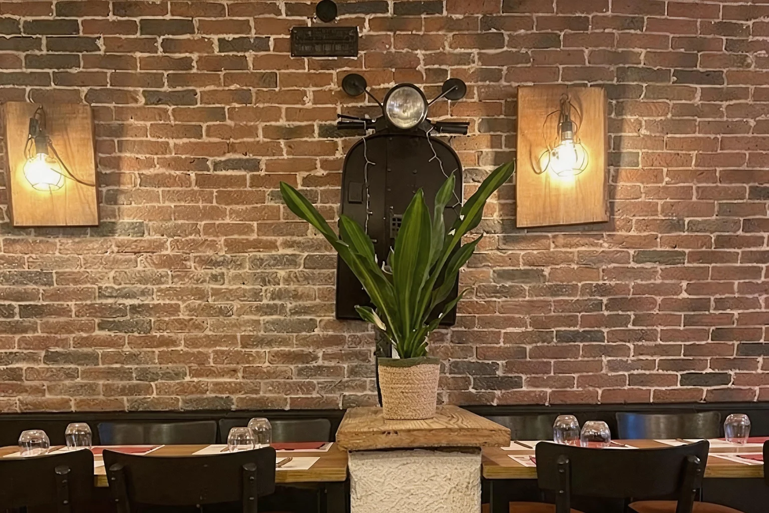 Dining room with brick wall, wall lamps, black scooter on the wall, plant and laid tables